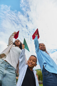 Sisters raising paper airplanes with younger brother in park with excited smiles. Creative learning, craft supplies, afterschool programs concept.