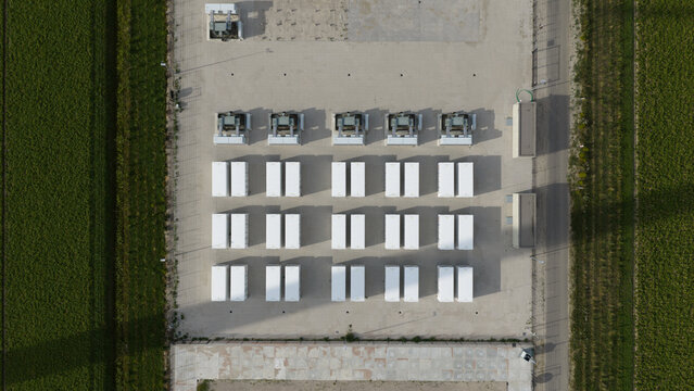 Aerial view of an energy storage facility with rows of white battery containers arranged in a grid on a gravel lot, surrounded by green agricultural fields in Dronten, Flevoland, Netherlands.