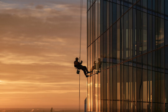 Industrial climber rappelling down a modern glass skyscraper at sunset.