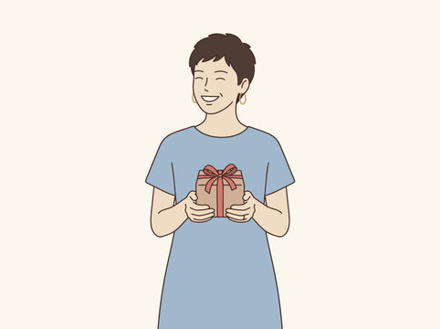 A cheerful young person with short dark hair wearing a blue shirt holds a small wrapped gift box with a red ribbon while smiling with closed eyes in a studio se