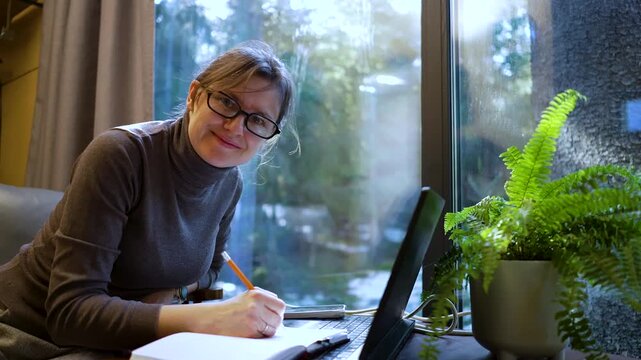 Freelancer lady types on laptop in serene home office. Focused woman professional creates content near window with natural light. Peaceful digital workspace for remote work.
