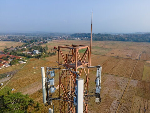 Aerial view of a telecommunications tower with antennas standing in a rural agricultural landscape with fields and trees in Patikraja, Central Java, Indonesia.