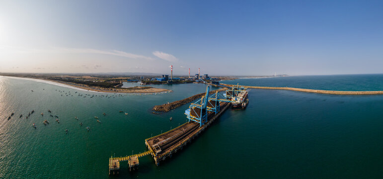 Aerial view of the power plant and coal terminal with industrial cranes, a cargo ship at the pier, and small fishing boats in the coastal waters in Cilacap, Central Java, Indonesia.