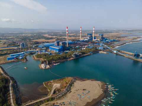 Aerial view of the coal-fired power plant with tall chimneys and industrial buildings along the coastline, with small fishing boats moored in the bay in Cilacap, Central Java, Indonesia.