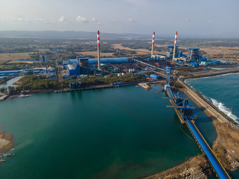Aerial view of the PLTU Cilacap power plant with its tall chimneys, industrial facilities, and coal conveyor belt extending into the harbor in Cilacap, Central Java, Indonesia.