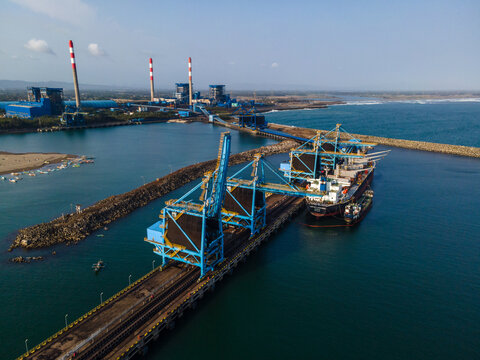 Aerial view of a large cargo ship being loaded with coal at an industrial terminal jetty, featuring blue cranes and power plant chimneys in the background, Cilacap, Central Java, Indonesia.