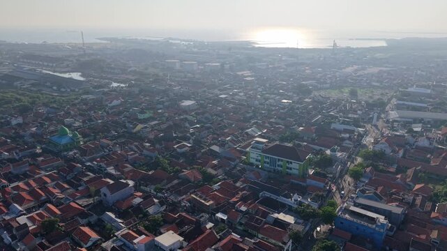 Sunlit aerial panorama of Probolinggo town with rooftops, mosque, industrial tanks, and distant coastline.