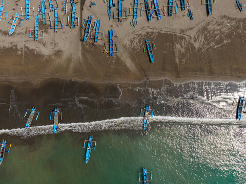 Aerial view of traditional blue fishing boats resting on the dark sandy beach and floating in the turquoise coastal waters of Cilacap, Central Java, Indonesia.