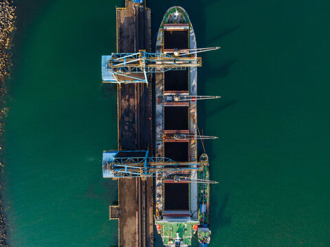 Aerial view of a large cargo ship being loaded with coal by industrial cranes at a port terminal in Cilacap, Central Java, Indonesia.