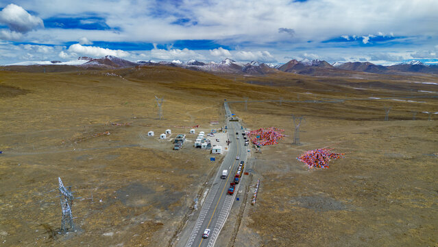 Aerial view of Gyatso La Pass on the Friendship Highway, featuring a road with vehicles, prayer flags, power lines, and arid terrain against snow-capped mountains in Shigatse, Tibet, China.