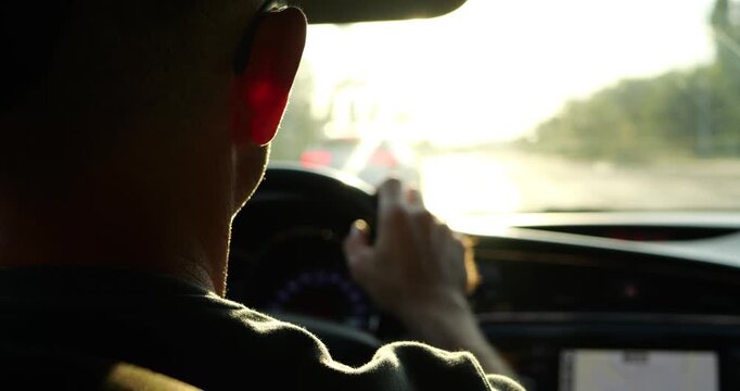 Male driver navigates sunlit road from car interior. Hand moves by steering wheel during commute, with navigation screen glowing. Warm light creates focus for road safety, mobility, and driving