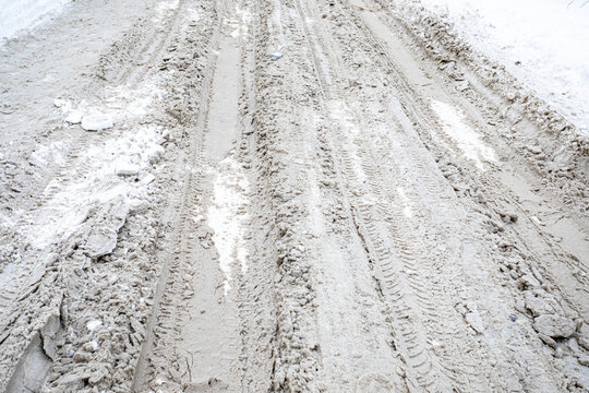 Parallel tire tracks cut through slushy snow and exposed mud, creating a rough winter road texture with uneven ridges, compacted ice, and pale white ground extending toward the background