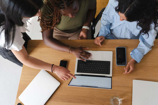 Three diverse female colleagues collaborate on a project inside a bright office. They lean over a wooden desk together, pointing at a laptop screen and discussing ideas.