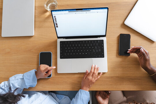 Two women sit together at a wooden table, browsing clothing items on a laptop computer. They discuss fashion trends while using multiple smartphones in a modern workspace.