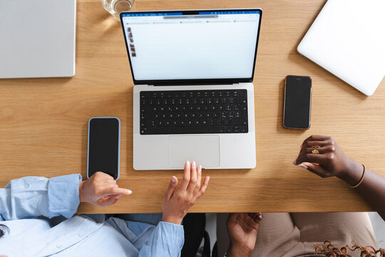 Two diverse women collaborate at a wooden desk while browsing an online fashion store on a laptop. Multiple devices surround them as they work together on a project.