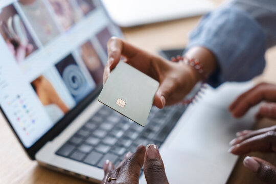 A person sitting at a desk with a laptop holds up a credit card to make an online purchase. Another person reaches out to help with the secure transaction.