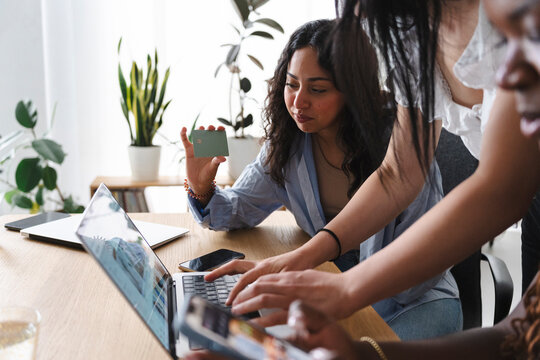 Three young diverse women collaborate in a bright office. One woman holds a green payment card while another types on a laptop computer, managing online finances together.