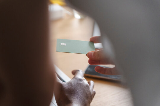 A person sitting at a desk holds up a green payment card to show someone else in a bright office space. They are managing personal finances during a workday.