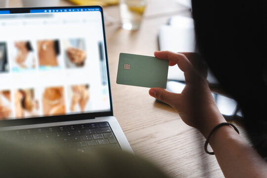 A person sits at a wooden desk with a laptop open to an online store, holding a green payment card in their hand while getting ready to purchase some new jewelry.