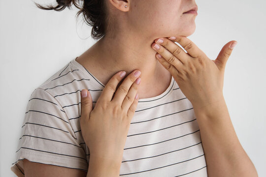 Woman with dark hair in bun, striped shirt palpating neck under jaw with both hands, close up, white wall, concept of swollen lymph nodes, thyroid self-examination, sore throat, endocrinology
