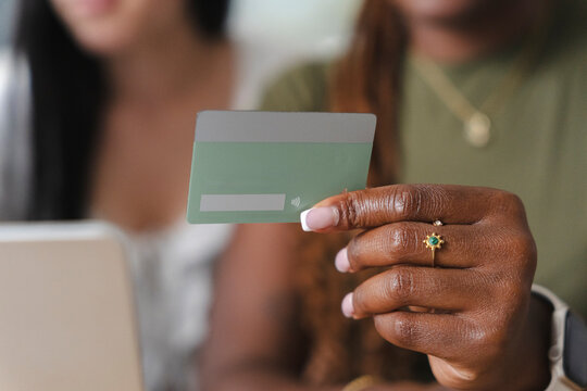 A Black woman holds a green credit card in her hand while making an online purchase on her laptop. Another person sits beside her in a modern brightly lit room.