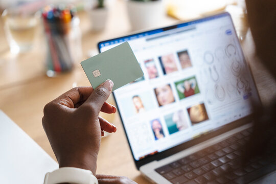 A person holds a green credit card in front of an open laptop computer. They are shopping online from home using a modern payment method to complete their digital purchase.