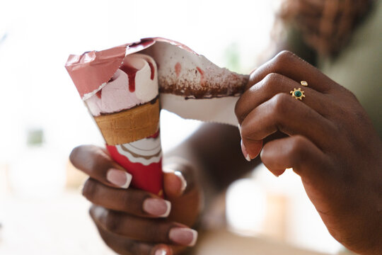 A person with dark skin and manicured fingernails carefully peels the paper wrapper off an ice cream cone with pink strawberry flavor, enjoying a sweet dessert.