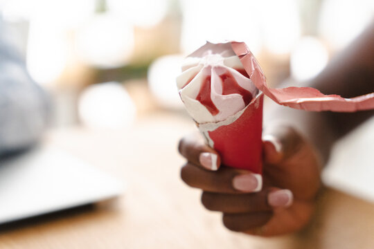 A person with dark skin holds a strawberry ice cream cone in a red paper wrapper, carefully peeling back the packaging to enjoy a sweet dessert during a sunny afternoon break.