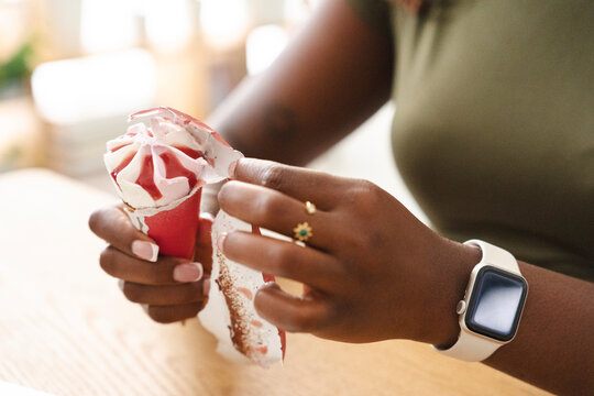 A Black woman unwraps a creamy red and white ice cream cone at a wooden table. She gently peels back the paper packaging to enjoy a sweet dessert during a relaxing break.
