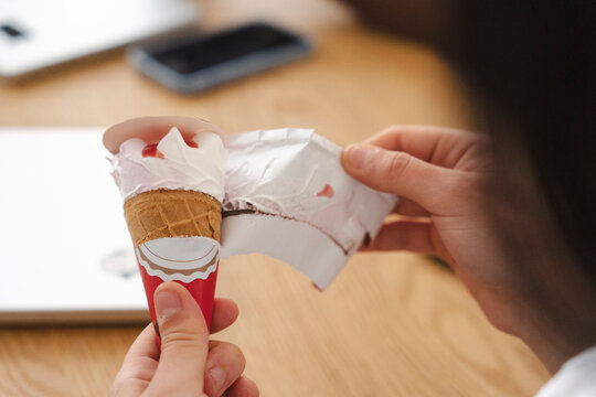 A person sitting at a wooden table is carefully unwrapping a delicious ice cream cone with vanilla and strawberry topping. Their hands peel away the paper.