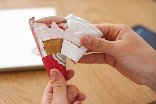 A person uses their hands to unwrap a packaged ice cream cone at a wooden table. The red and white wrapper is being peeled back to reveal the wafer cone inside.
