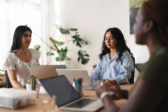 Three diverse women sit together around a wooden table in a brightly lit office space. They are focused on their laptops, collaborating on a shared work project.