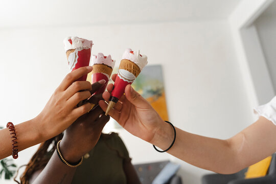 Three diverse friends hold their ice cream cones together in a celebratory toast indoors. They enjoy sharing a sweet treat during a casual gathering at home.