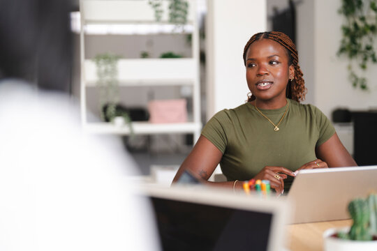 A young Black businesswoman with braided hair sits at a wooden desk in a bright office, talking with a colleague during a collaborative project meeting.