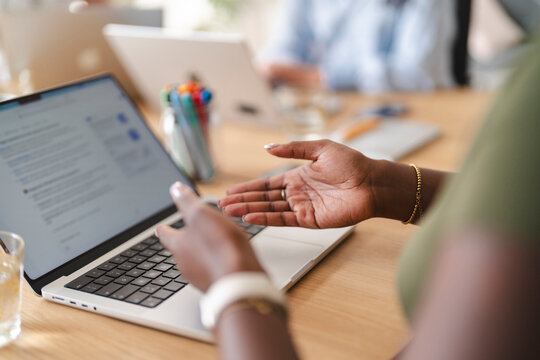 A Black businesswoman sits at a wooden office desk, gesturing with her hands while working on her laptop during a collaborative team meeting with colleagues.