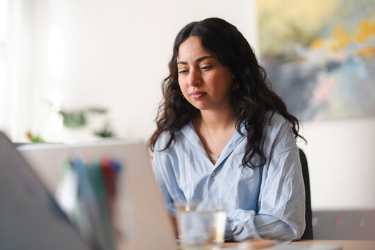 A young woman with dark hair sits at a wooden desk in a bright room. She is focused on her work, looking down at a digital tablet while typing or reading.