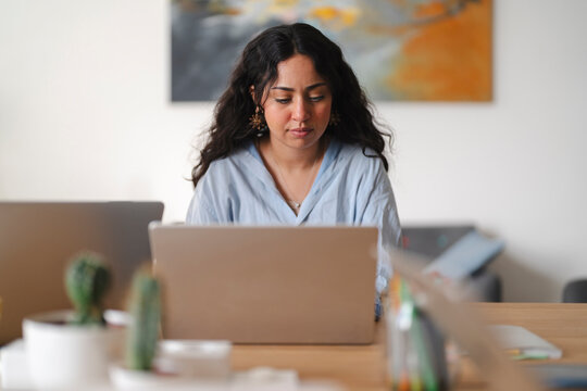A young woman with long dark curly hair is sitting at a wooden desk in a bright modern office, fully focused on typing and working on her laptop computer.