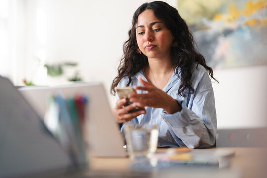 A young woman with dark curly hair sits at a desk in a bright room. She is focused on her smartphone while working from home, holding the device and typing a message.