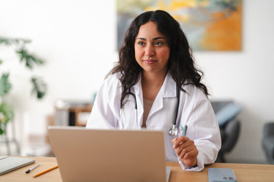 A young female doctor wearing a white coat and stethoscope sits at a wooden desk in her office, looking away from her laptop while thinking during a consultation.