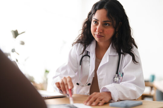 A young female doctor wearing a white coat and stethoscope sits at a wooden desk, pointing a red pen at documents while looking intently at her laptop during a consultation.
