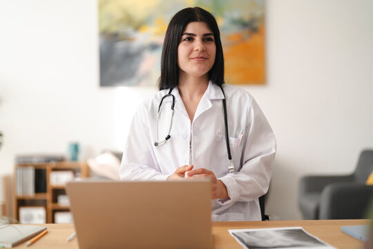 A young female doctor wearing a white coat and stethoscope sits at her office desk with a laptop. She smiles gently while having a consultation with a patient.
