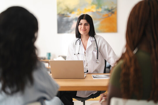 A female doctor sits at her desk with a laptop and consults with two women in her bright medical office. They discuss health options during a professional appointment.