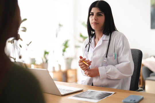 A female doctor wearing a white coat and stethoscope sits at her desk in a clinic, having a consultation with a patient and explaining medical results using a laptop.