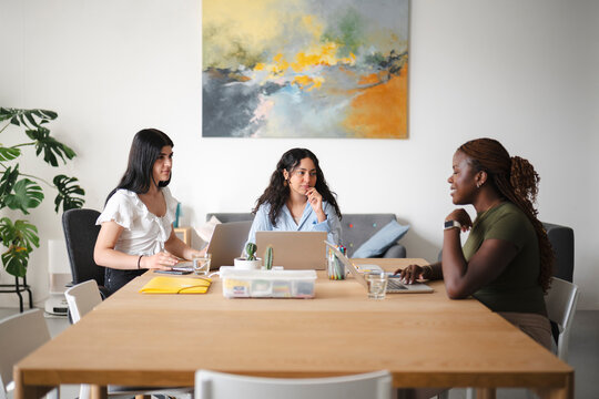 Three diverse women sit together at a wooden table in a modern office, collaborating on projects using portable computers and discussing ideas in a bright workspace.