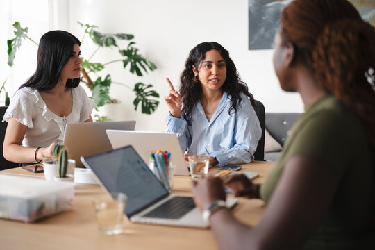 Three diverse women collaborate in a modern office space. They sit around a wooden table with laptops, actively discussing a project during a productive team meeting.