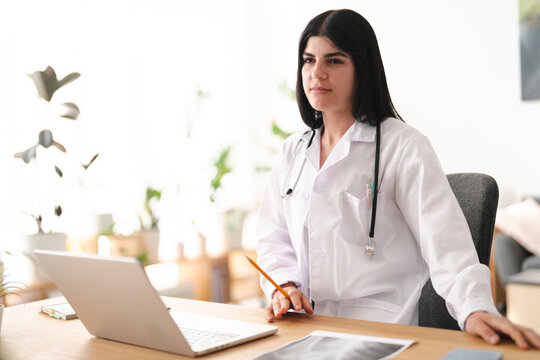 A young female doctor wearing a white lab coat and stethoscope sits at a wooden office desk. She holds a pencil while looking away thoughtfully, with a laptop nearby.