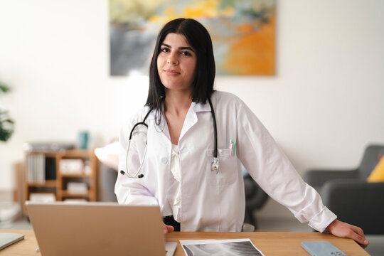 A young female doctor stands behind her office desk, looking directly at the camera with a confident expression. She wears a white coat and a stethoscope around her neck.