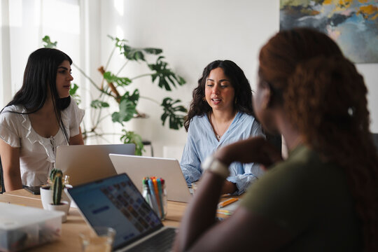 A diverse group of three female colleagues collaborate in a bright modern office. They sit together around a wooden table, working on laptops and discussing their project.