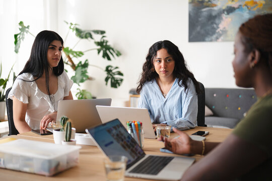 A diverse group of young female colleagues collaborate in a bright modern office. They sit around a wooden table with laptops, discussing a business project and sharing ideas.