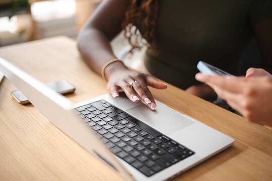 A Black woman works at a wooden desk, using the trackpad on a silver laptop. Another person sits nearby, holding a smartphone while collaborating on a project together.